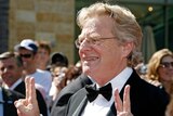 Jerry Springer shows two finger peace signs with both hands as he smiles while wearing a tuxedo.