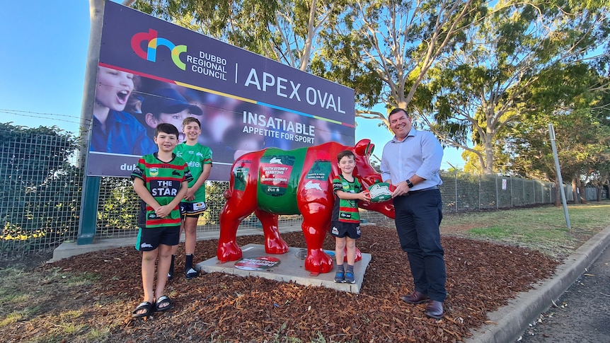 A man and three children stand in front of a sign for a football field and a red and and green painted rhinoceros statue.