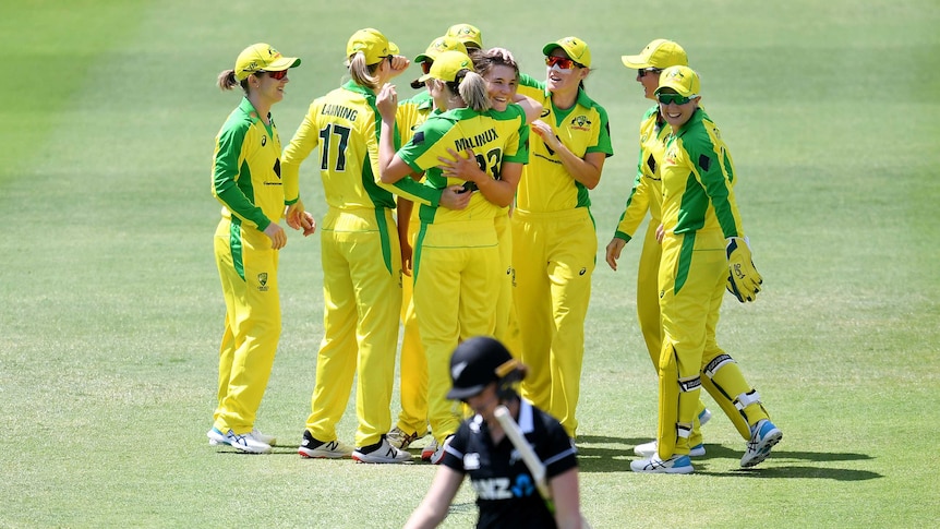Australian players hug in a large group as a New Zealand player wearing black walks in the foreground