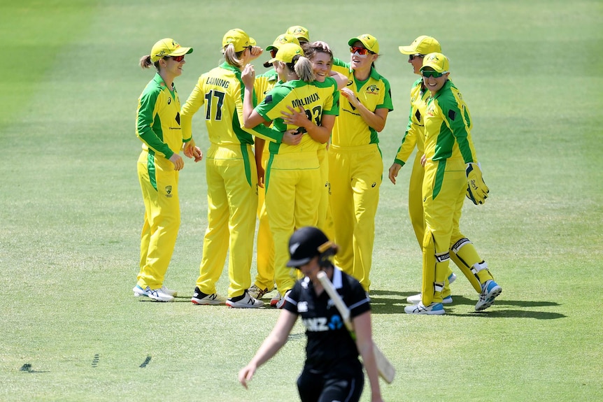 Australian players hug in a large group as a New Zealand player wearing black walks in the foreground