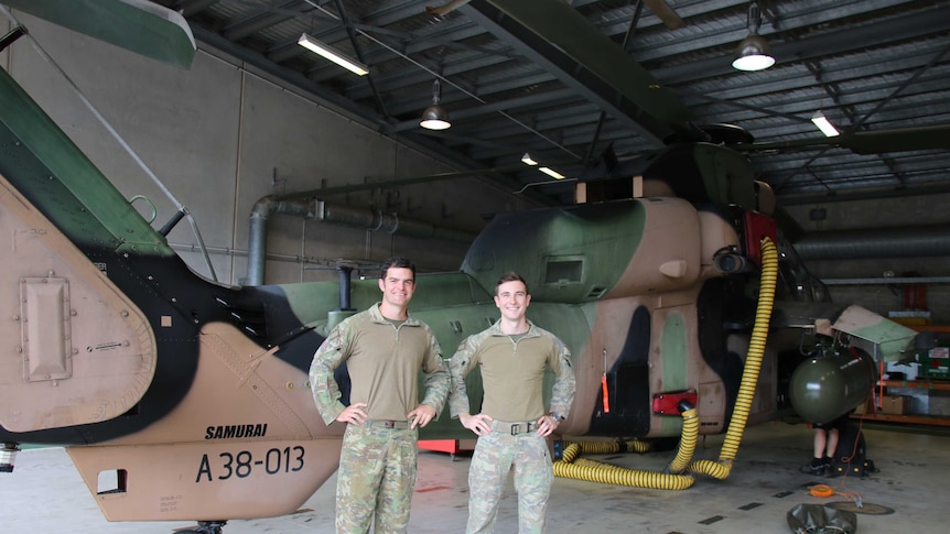 Two young men in army camouflage standing before a Tiger helicopter, their hands on their hips