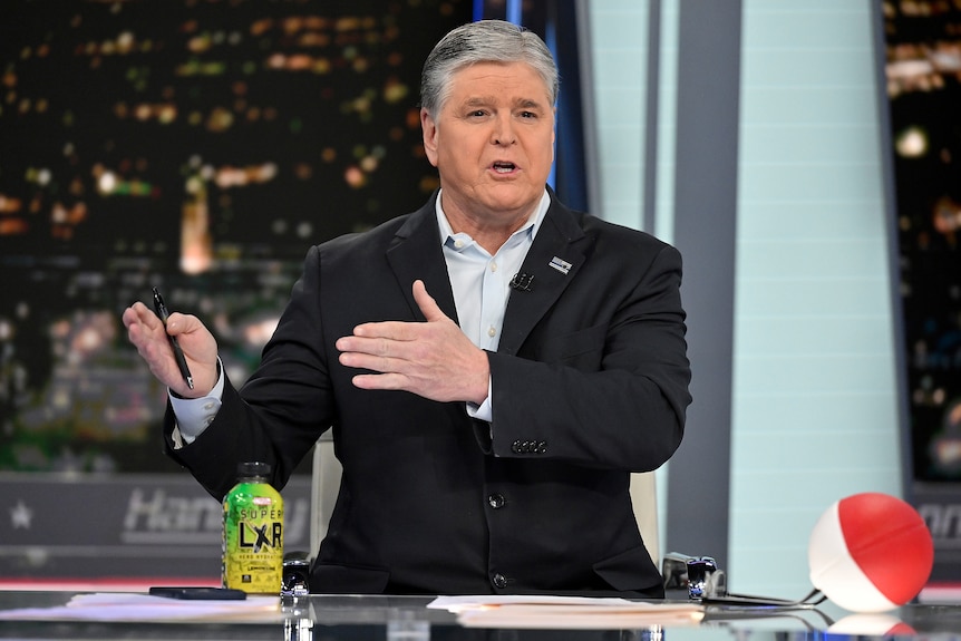 Sean Hannity gestures while sitting at a news desk with a bottle of energy drink and holding a pen