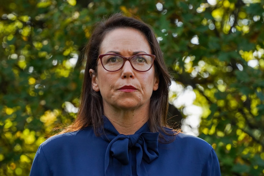 Jaclyn Symes, dressed in a blue blouse, appears determined as she stands underneath a leafy tree on a sunny day.