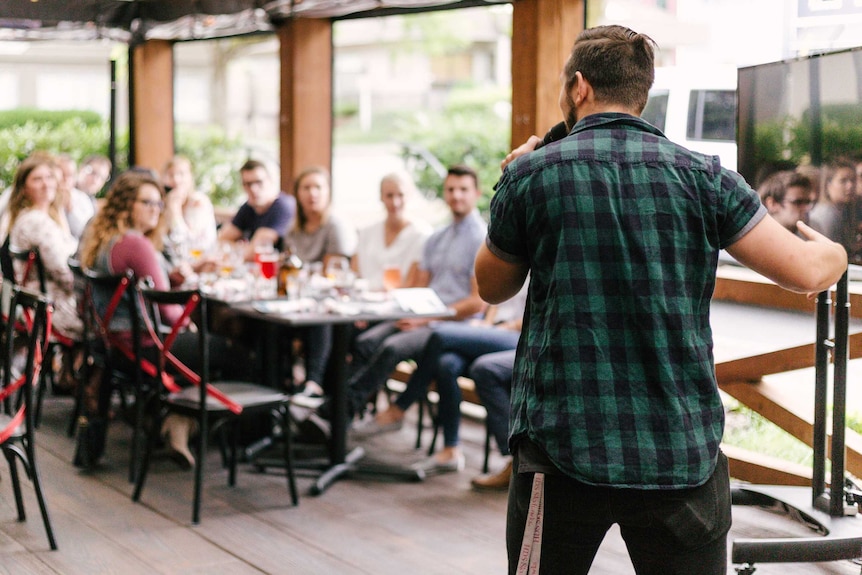 Before a blurred, seated crowd at a long table, a man in short-sleeved chequered shirt is seen from behind holding a microphone.