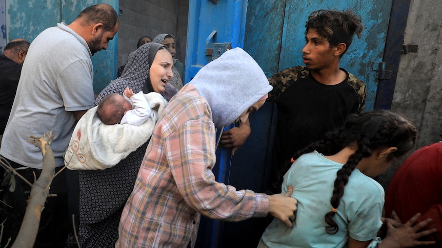 Five people are pictured, they look scared. One woman is holding a baby as she walks away with the group.