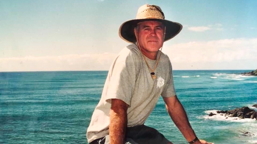 An man wearing a straw hat and boardshorts sits on a wooden rail on the coast.