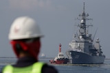 A tug boat pushes a large grey ship in water as a worker in a hard hat watches on from afar