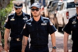 A man in NT Police uniform walking down the street, on a sunny day.