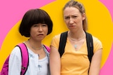 Two women pretending to be teenage girls stand awkwardly wearing t-shirts and backpacks, cut out against a pink, yellow backdrop