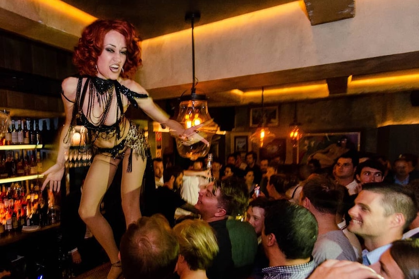 A burlesque dancer stands on a bar with a crowd of people watching