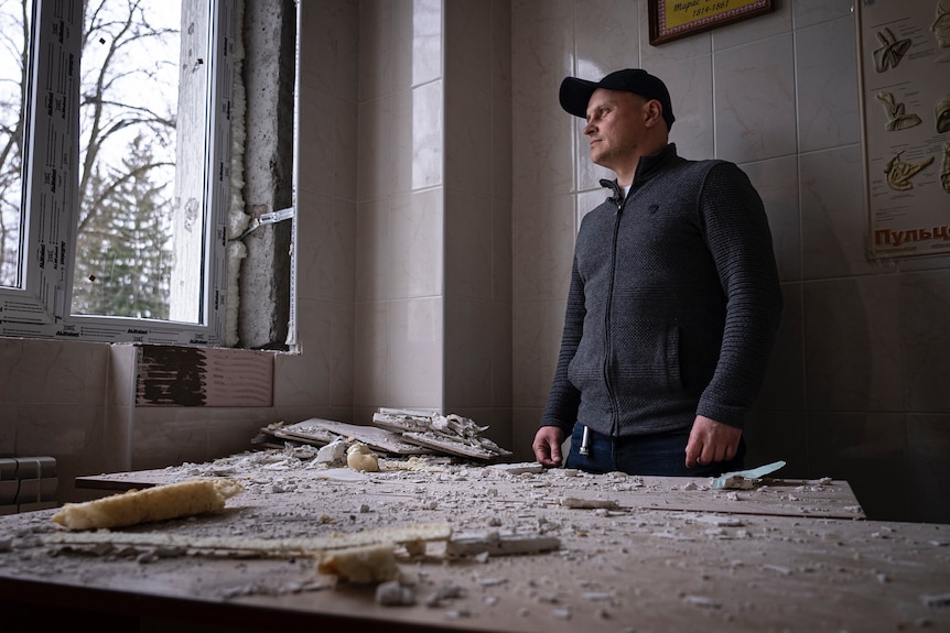 A man in a zip-up jumper and baseball cap looks out the window of a damaged hospital room