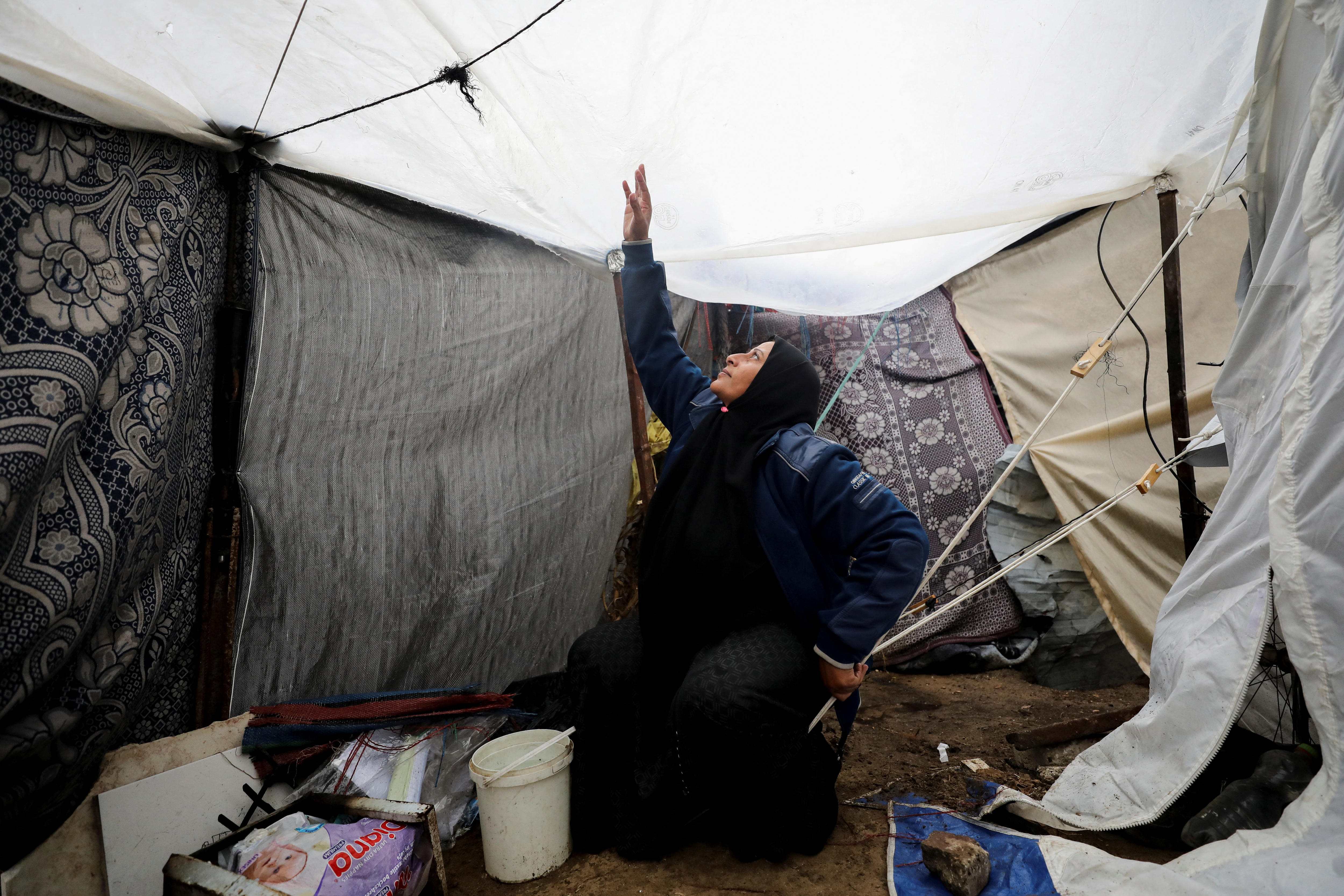 A middle-aged woman in a black hijab and long sleeves reaches up to the drooping roof of a white tent, her fingers outstretched
