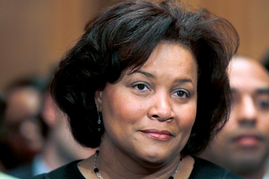 A black woman with short hair stands in a courtroom, staring neutrally ahead.