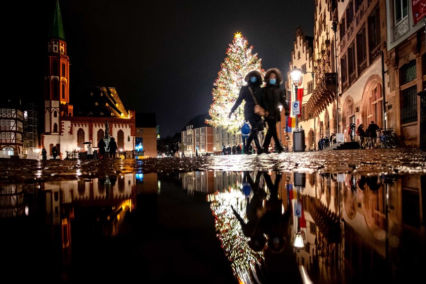 Two women in face masks and coats walk past a Christmas tree in a town square