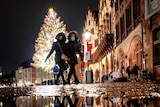 Two women wearing face masks down a street a night with a brightly lit Christmas tree in the background.