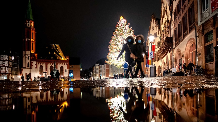 Two women wearing face masks down a street a night with a brightly lit Christmas tree in the background.