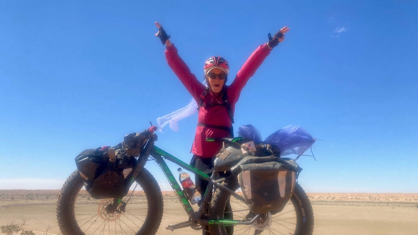 Woman in a bike helmet, pink zip up jacket and sunglasses, standing in the desert front of push bike, with arms in the air