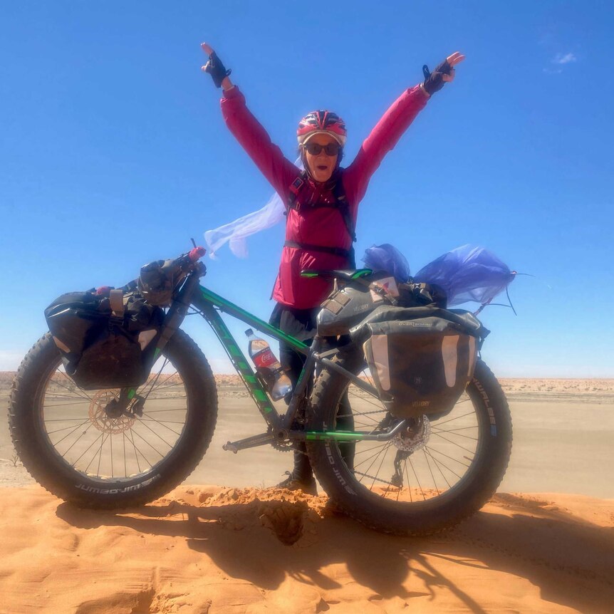 Woman in a bike helmet, pink zip up jacket and sunglasses, standing in the desert front of push bike, with arms in the air