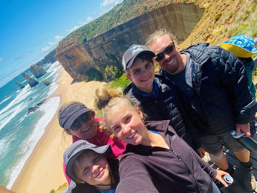 A family of five smile at the camera for a selfie with a coastline in the background.