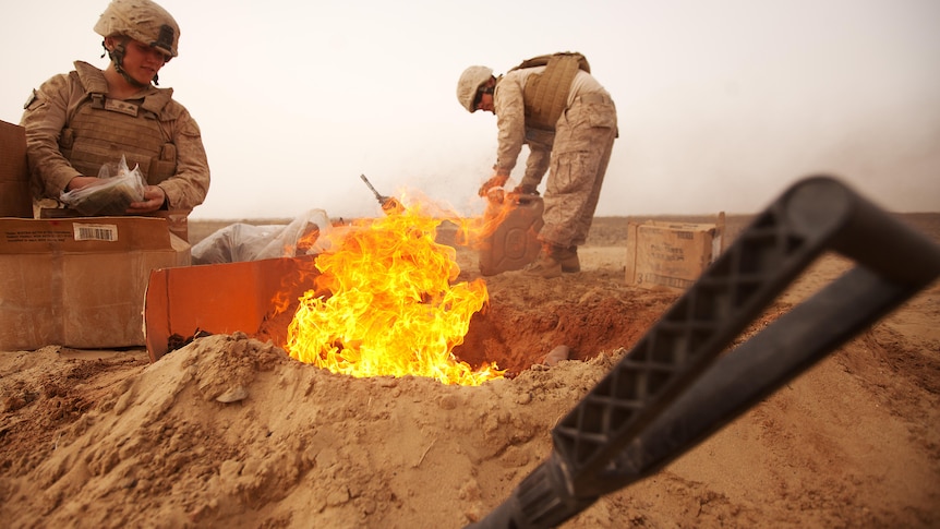 Two military personnel stand next to a burn pit in Afghanistan.