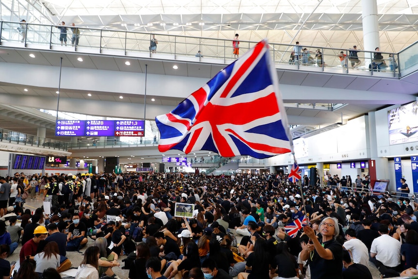 From a high angle, you look down on a large crowd of black t-shirt-clad protesters as a woman at its centre waves a British flag
