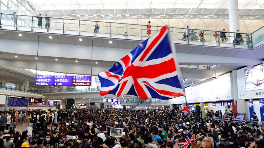 From a high angle, you look down on a large crowd of black t-shirt-clad protesters as a woman at its centre waves a British flag