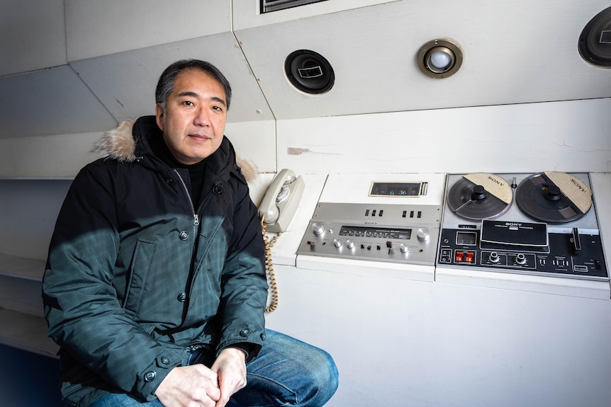 一名身穿毛边夹克的日本男子坐在接近 20 世纪中期的技术舱内