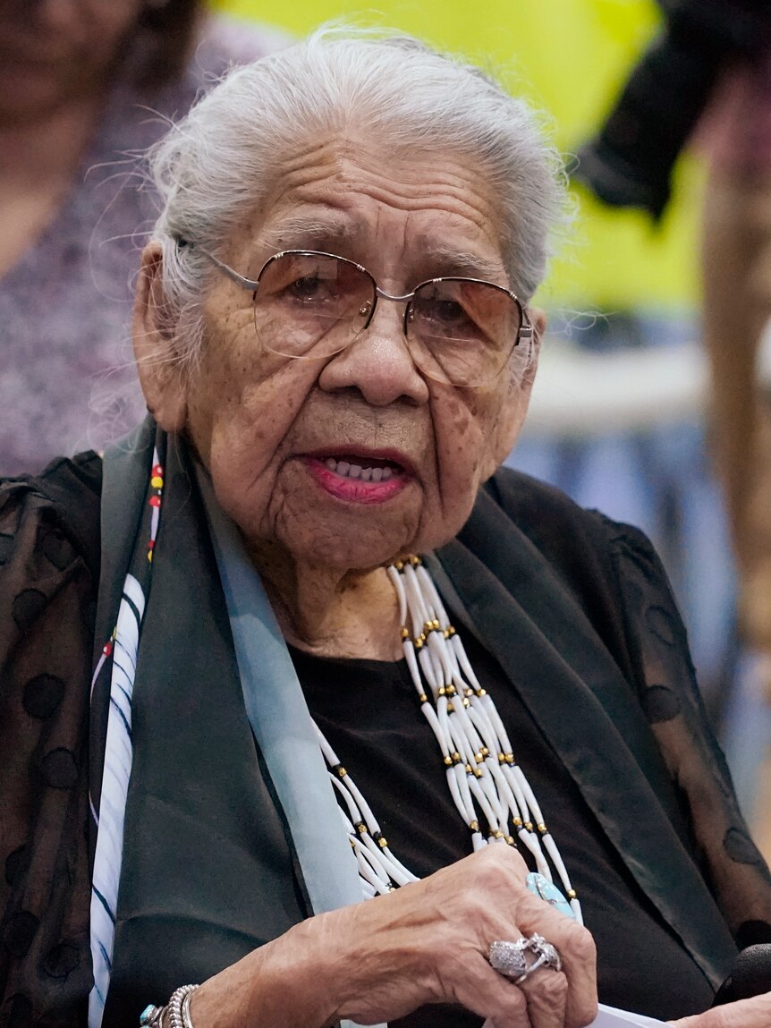 An elderly lady wearing glasses and a beaded necklace speaks while sitting a wheelchair.