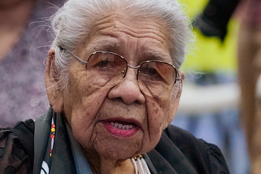 An elderly lady wearing glasses and a beaded necklace speaks while sitting a wheelchair.