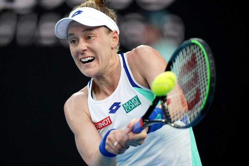 A tennis player braces as she swings her racquet at the ball for a two-handed backhand.