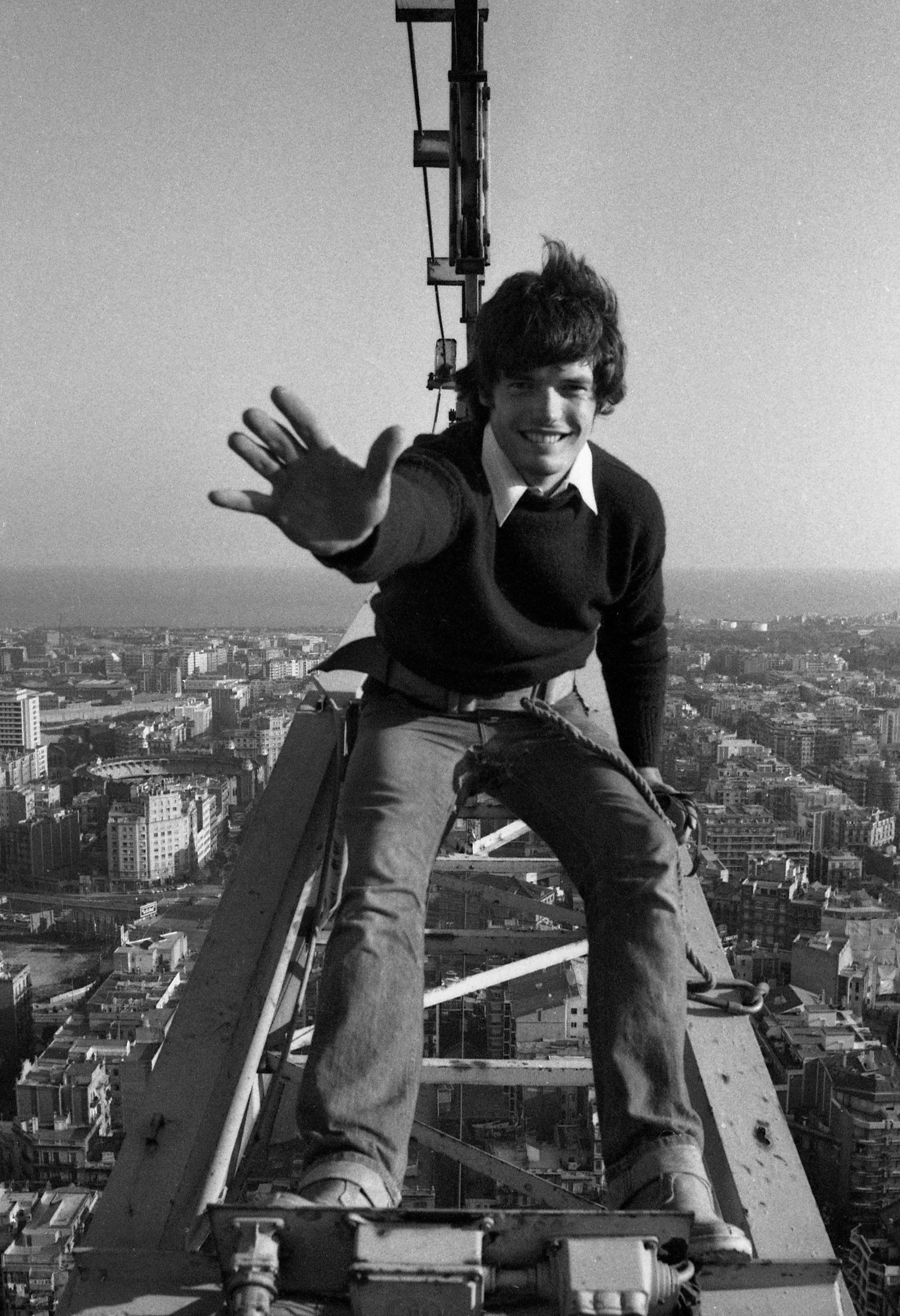 A black and white image of a young man standing on top of a crane reaching forward with his hand and smiling