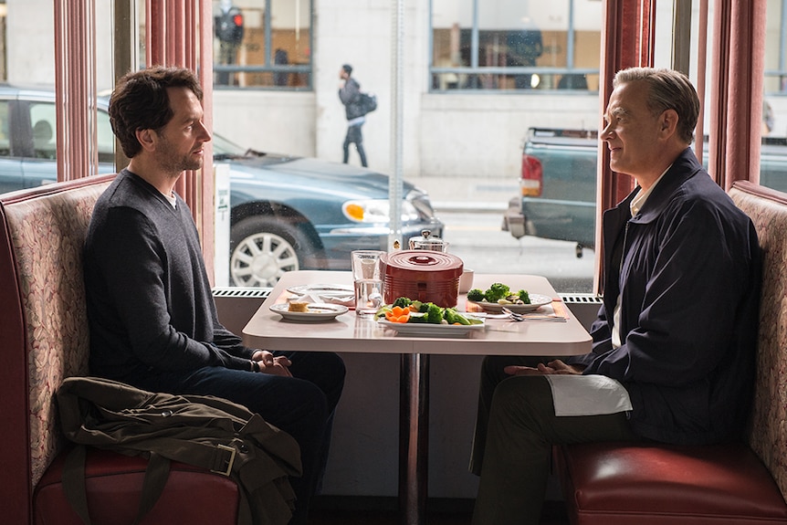 Two men, one older and one middle aged, sit opposite each other in diner booth next to street level window with food on table.