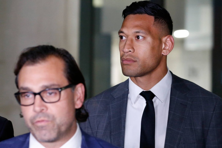 Israel Folau looks to one side wearing a dark grey suit with a black tie and white shirt, standing behind a man in glasses