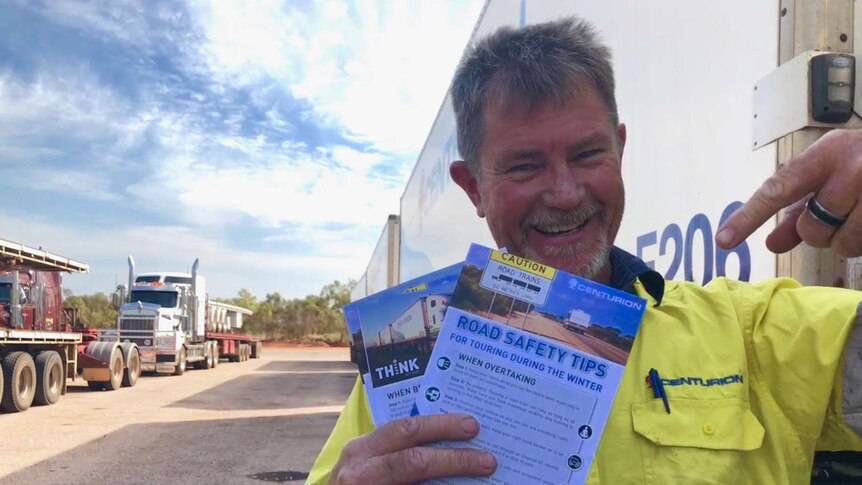 A truck driver standing in front of a triple road train at a roadhouse in Broome, holding safety brochures
