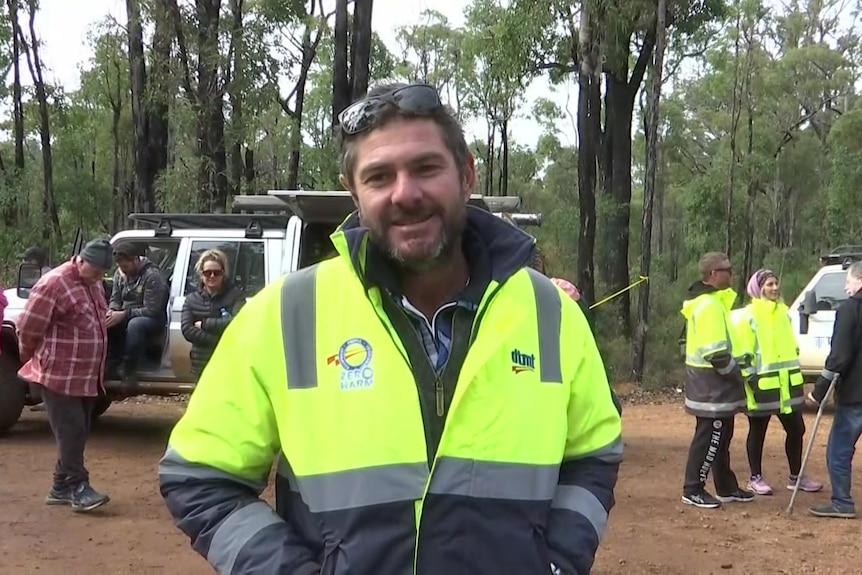 A man wearing a hi-vis jumper in bushland, with people and a car in the background. 