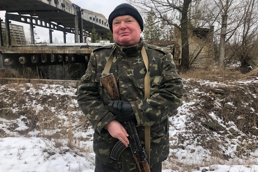 A man in a beanie and camouflage outfit cradles an assault rifle.