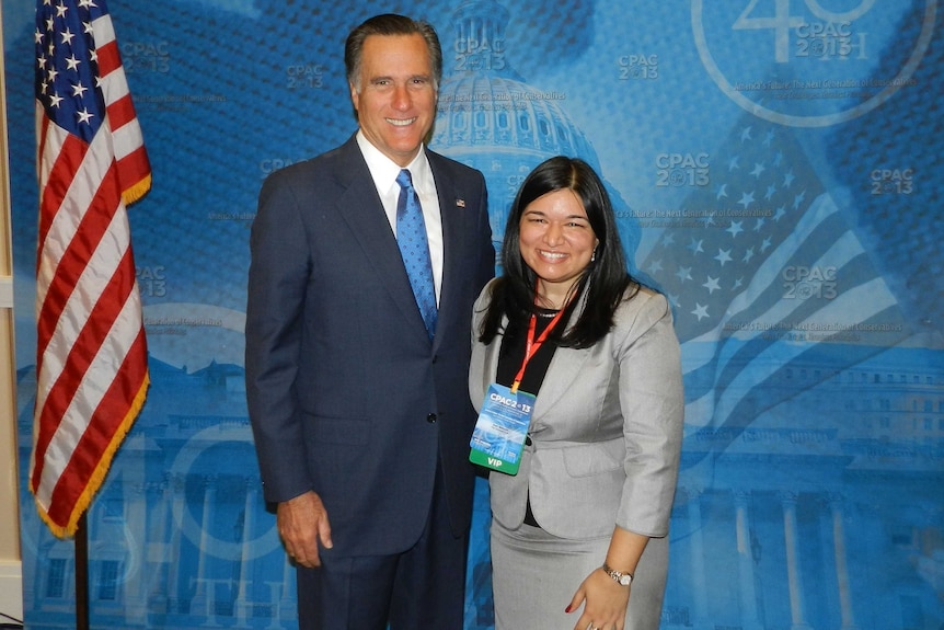 A man and woman pose for a photo next to a flag against a blue background.