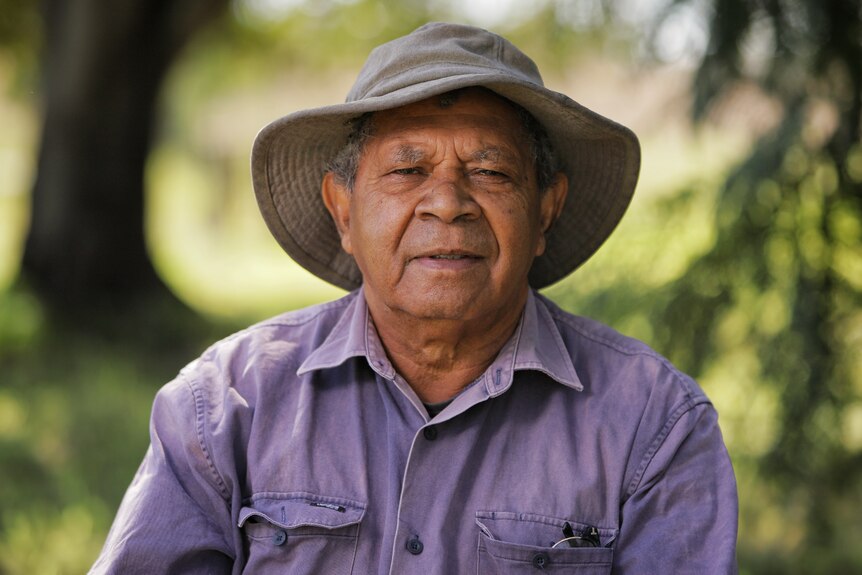 Indigenous man wearing a purple shirt and hat, sitting in the bush.