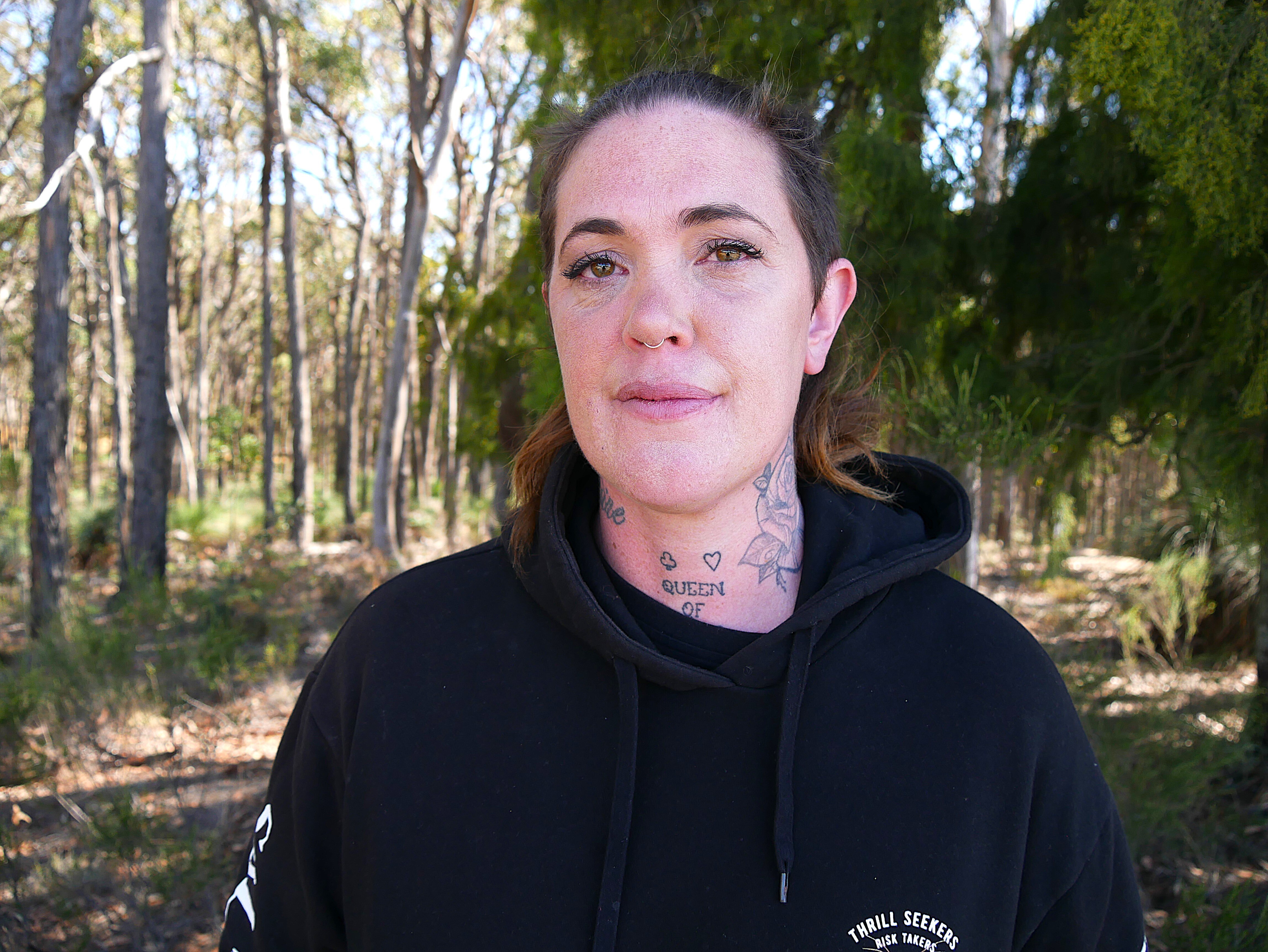 a woman with tattoos and long brown hair tied back in a ponytail is standing in bushland staring at the camera 
