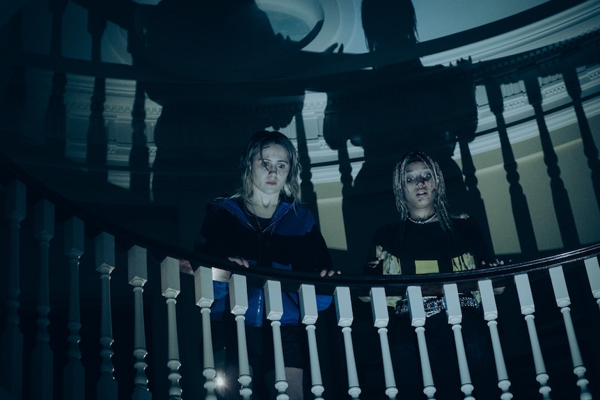 Low-angle photo of two women in their twenties at the top of a flight of stairs, looking dismayed by bloodied faces. 