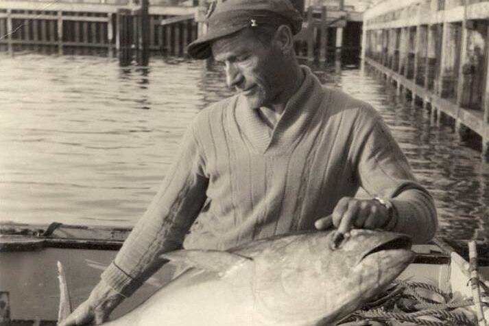 Black and white photo of man in cap holding large tuna fish on back of boat, at jetty