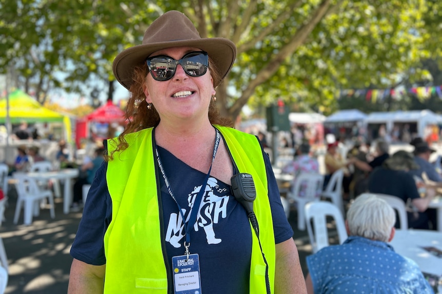 A woman in an akubra and a fluorescent yellow vest smiles at an outdoor festival.