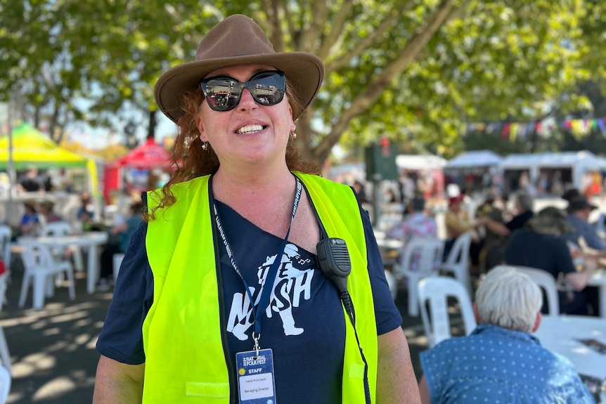 Una mujer con una akubra y un chaleco amarillo fluorescente sonríe en un festival al aire libre.