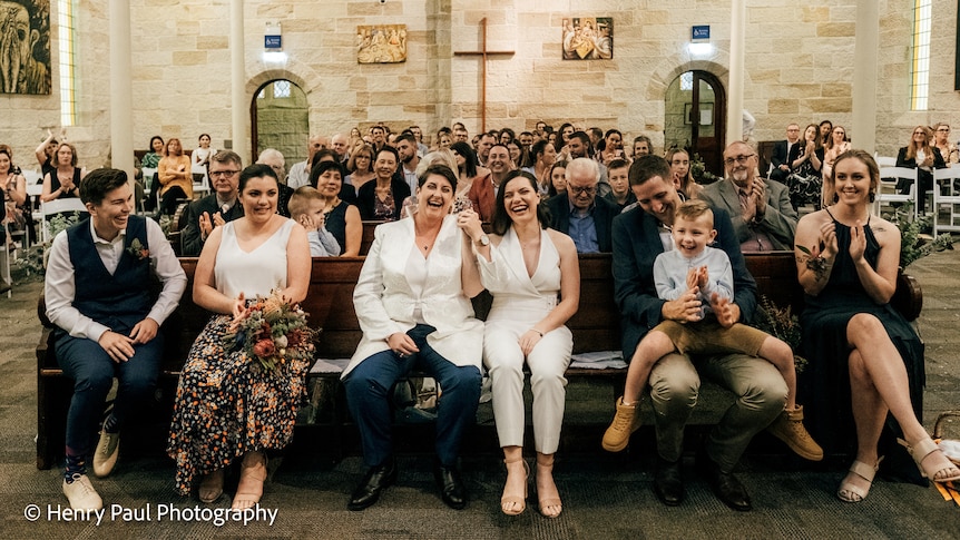 Two women on their wedding day sitting in a church surrounded by loved ones. 