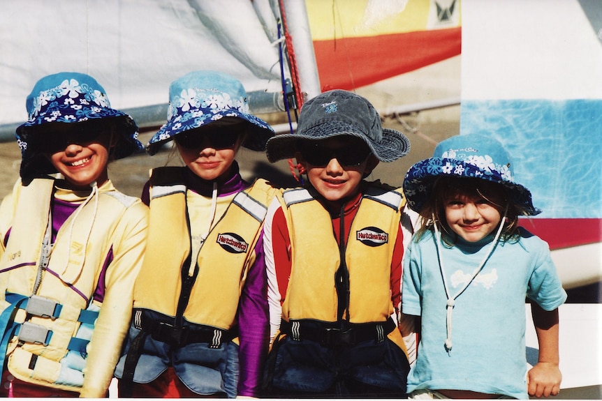 Four young kids, including Jessica Watson, wearing life jackets and standing in front of a sail boat, ocean behind.
