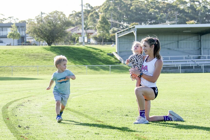 Amy O'Halloran watches her son Henry (3) run on a grassed oval, while she kneels and holds her daughter Jemima (1).