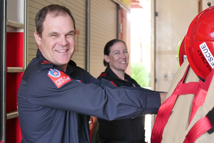 A man and woman wearing blue Fire and Rescue uniforms stand smiling at the fire station, reaching for their jackets.