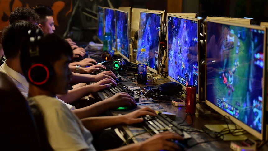 Young Chinese men sit in a row wearing headphones and playing online games in a darkened room.