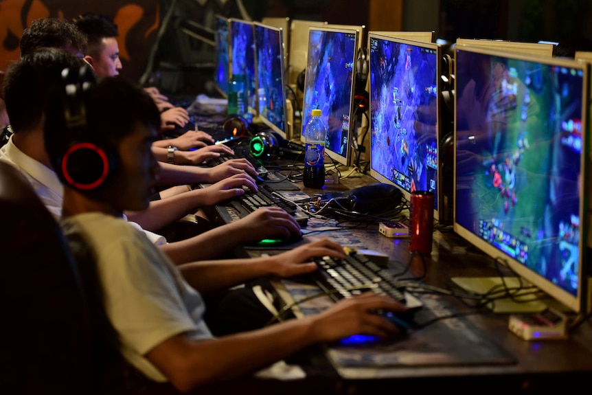 Young Chinese men sit in a row wearing headphones and playing online games in a darkened room.