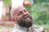 Man with beard and beige shirt laughs with eyes closed. 